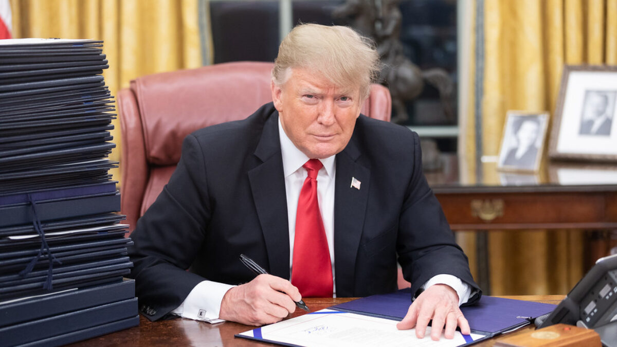 A straight-faced Donald Trump signs a document in the Oval Office