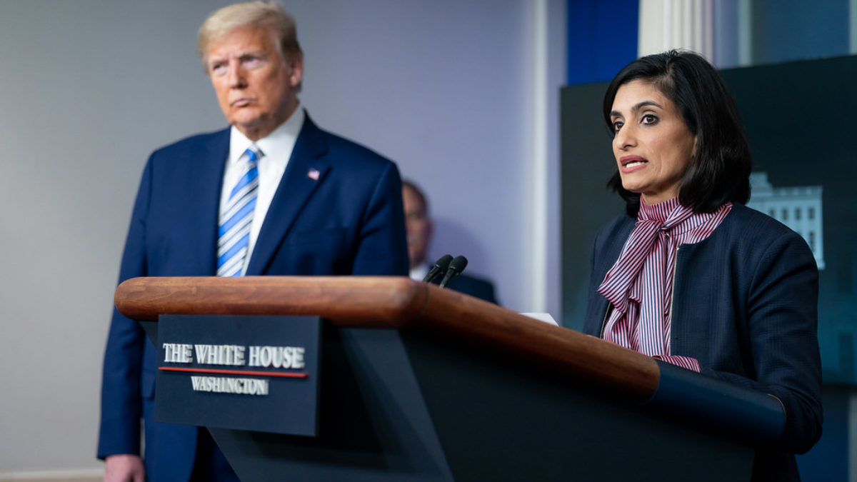 CMS Administrator Veema Serma speaks at a podium at a press briefing while Donald Trump looks on
