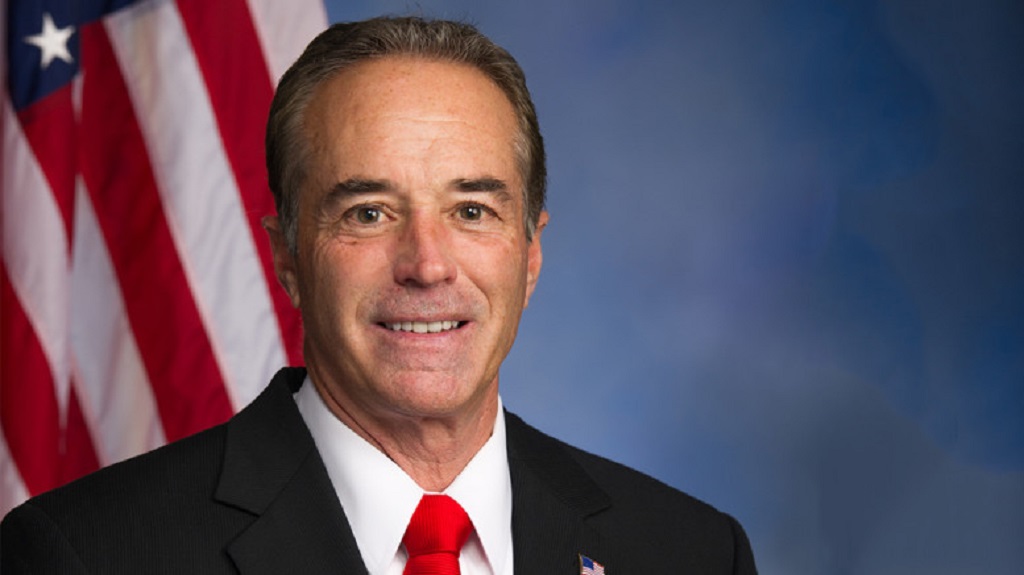 Chris Collins sits in front of a blue backdrop with the American Flag