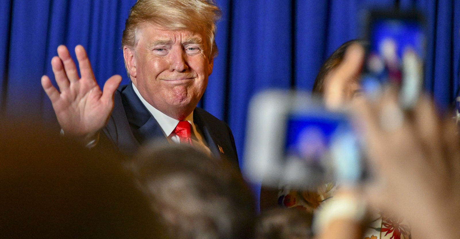 President Trump waves at a crowd in front of a blue backdrop