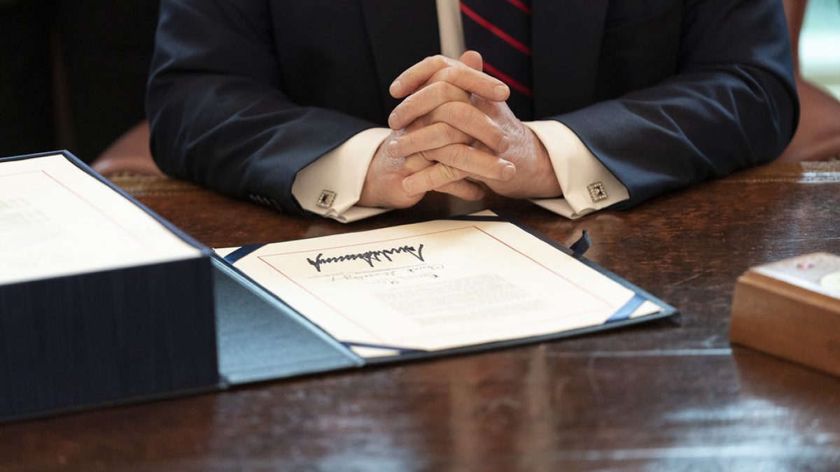 Document with Trump's signature in the foreground with Donald Trump's crossed hands in the background