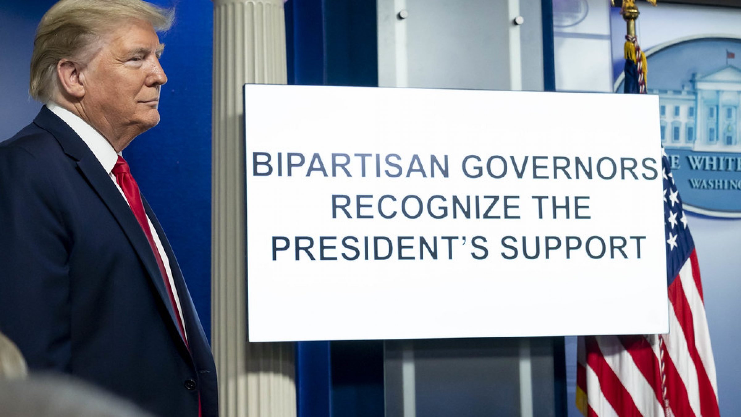 Trump smirks aside a sign that reads "Bipartisan Governors Recognize the President's Support" in all capital letters in the White House Press Briefing Room.