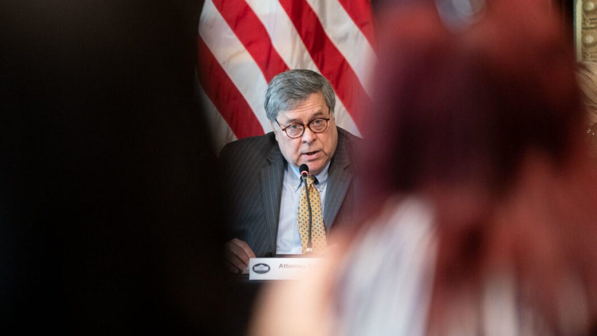 Bill Barr speaks while seated in front of an American flag