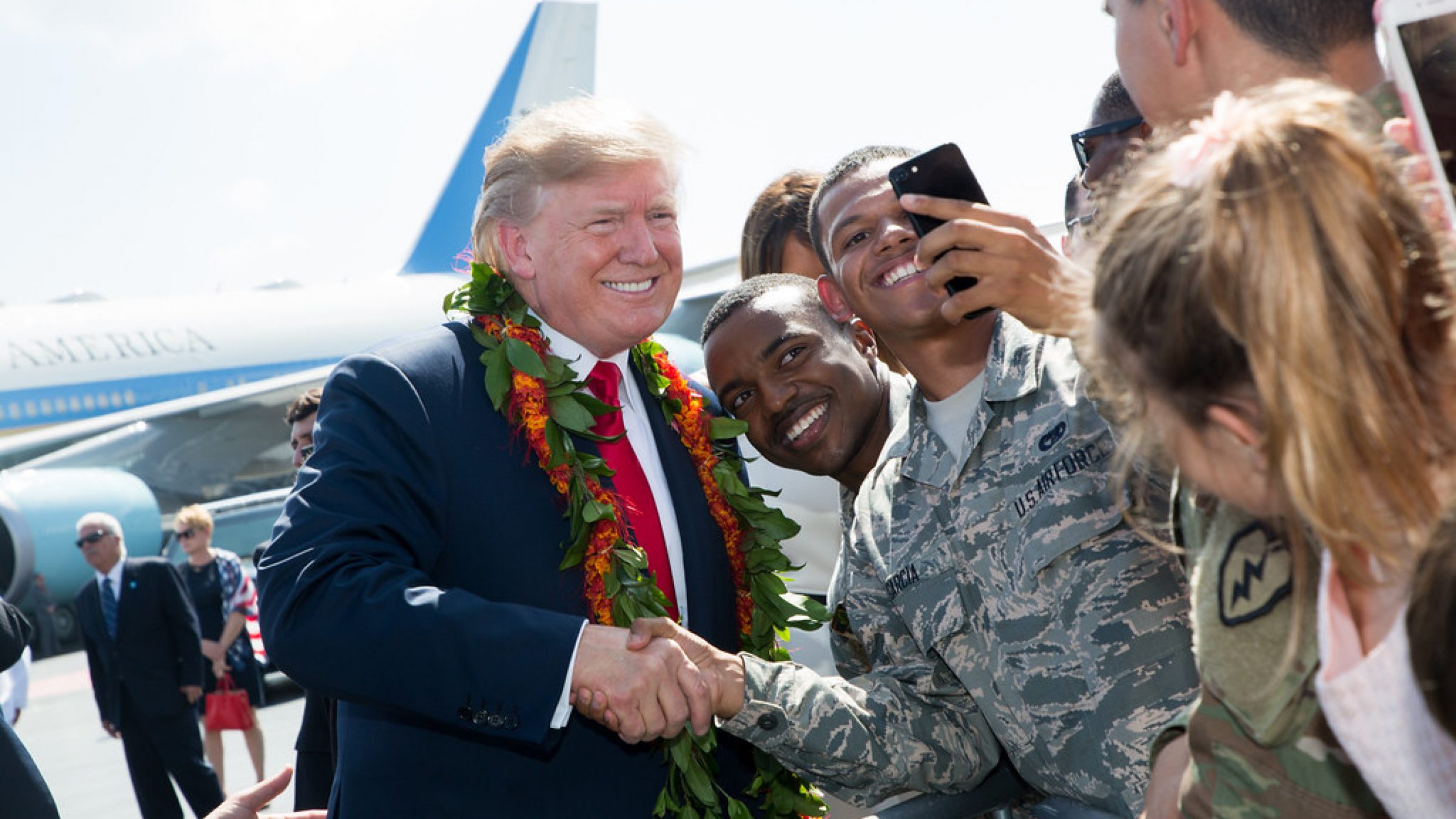 Donald Trump, wearing a lei, shakes hands and smiles for a selfie with a member of the US Air Force